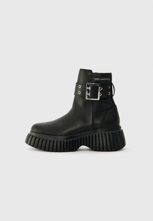 Black leather ankle boots with a thick ribbed sole. Features a wide ankle strap with a silver buckle and eyelets. Minimal branding at the back.