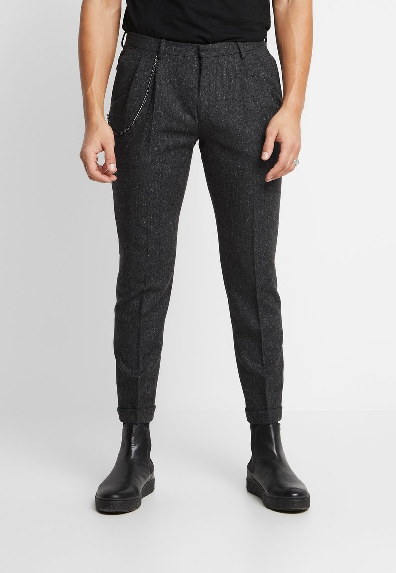 Donkergrijze op maat gemaakte broek van een gestructureerde stof, slim fit, opgerolde boorden en een zilveren kettingaccent. G gedragen met zwarte enkelboots.