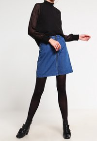 Blouse noire à pois avec manches transparentes, col montant et bordure à volants, associée  à une jupe en denim bleu à fermeture éclair sur le devant et des bottines noires.