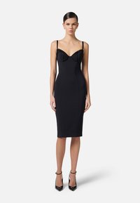 Abito nero aderente con scollatura a cuore, sottili spalline e coppe strutturate. Tessuto liscio, lunghezza al ginocchio, abbinato a tacchi neri.