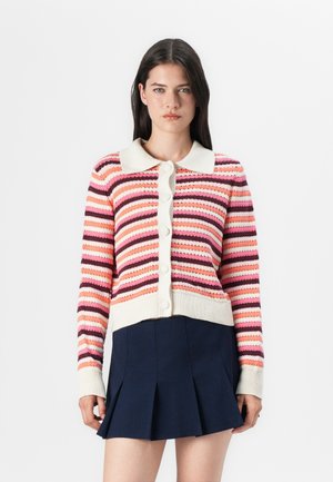 En ung kvinne står mot en nøytral bakgrunn, iført en stripete genser i rosa og hvite nyanser, kombinert med et marineblått plisserte skjørt.
