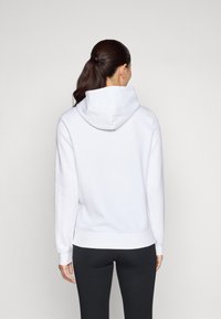 Felpa bianca aderente con un grande cappuccio e polsini a coste, realizzata in materiale morbido. Abbinata a leggins neri, vista da dietro.