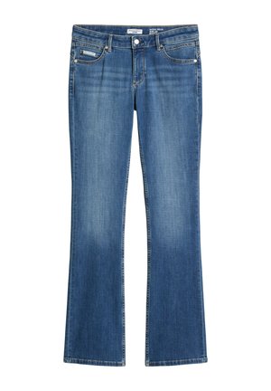 Junge Person trägt ein schwarzes langärmeliges Shirt, blaue Jeans und dunkle Schuhe und steht vor einem schlichten hellgrauen Hintergrund.