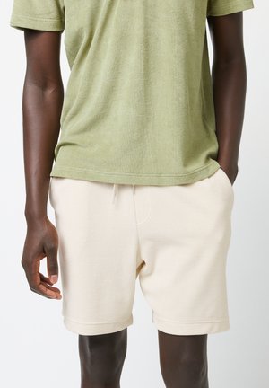 Camicia a maniche corte color verde oliva realizzata in tessuto morbido, abbinata a pantaloni corti color crema con cordoncino. Texture liscia, vestibilità morbida.