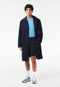 Abrigo largo azul marino, camiseta turquesa, pantalones cortos oscuros y zapatillas multicolores. Presenta botones y el logo de Lacoste en la camiseta y en las zapatillas.