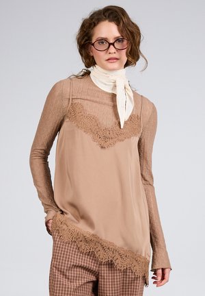 Kvinde iført brun blonder langærmet top, beige silkebluse med blondekant, ternede bukser, hvidt tørklæde og runde briller mod ensfarvet baggrund.