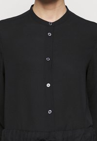 Blouse noire avec col mandarin, dotée de fermetures à boutons et d'une taille élastique. Tissu lisse avec une texture unie.