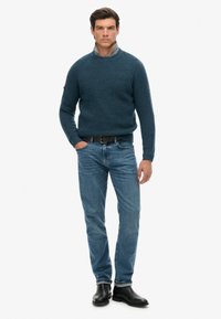 Blauer gestrickter Pullover mit Rippstruktur, runder Ausschnitt, kombiniert mit hellblauen Jeans und schwarzen Lederstiefeln, die Hände in den Taschen.