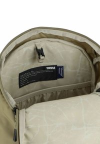 Intérieur de sac à dos Thule couleur crème avec une doublure lisse et texturée, une poche zippée et une étiquette présentant les détails de la marque et des matériaux.
