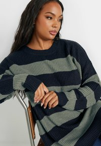 Randig stickad tröja med mörkblå och olivgröna linjer, ribbad textur, oversized passform och långa ärmar. Bäraren vilar händerna på en stol.