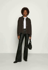 Μαύρο oversized πουκάμισο-τζάκετ, λευκή μπλούζα, μαύρα παντελόνια σε φάρδος, μαύρη τσάντα ώμου με πλεχτό λουρί, φορεμένα από ένα άτομο με σγουρά μαλλιά.