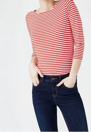 Personne portant un haut à manches longues rayé rouge et blanc, rentré dans un jean bleu foncé, les mains dans les poches avant, sur un fond uni.