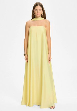 Frau trägt ein bodenlanges, trägerloses, blassgelbes Kleid mit passendem Halstuch, goldenem Armband und Ohrringen vor weißem Hintergrund.