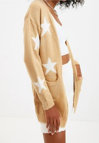 Lång beige kofta med vita stjärnmönster, öppen front, två fickor, gjord av mjukt material, med lösa ärmar och avslappnad passform.