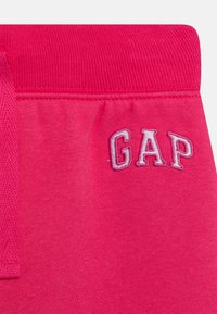Jasnoružové tepláky majú hladký povrch s elastickým pásom a vyšívaným logom "GAP" svetlo šedou farbou.