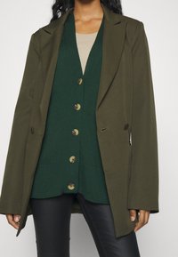 Olivgrön blazer lagd över en mörkgrön knappad cardigan med stora knappar och texturerad stickning. Bärs över slanka svarta byxor.