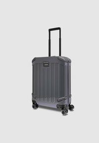 Valise trolley rigide en gris foncé, avec des rainures verticales, une poignée télescopique, quatre roues pivotantes et des accents en caoutchouc.