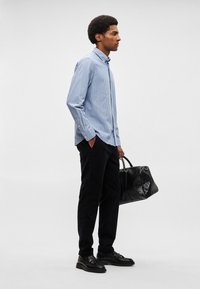 Ανοιχτό μπλε ριγέ πουκάμισο με κουμπιά, μαύρο παντελόνι κοτλέ και μαύρη υφασμάτινη τσάντα. Το μοντέλο στέκεται στο πλάι, παρουσιάζοντας την εμφάνιση και τα αξεσουάρ.