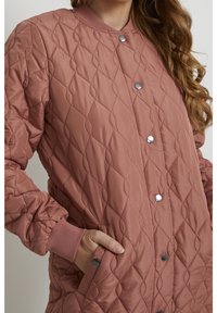 Femme portant une veste matelassée rose poussiéreux avec des boutons-pression et la main droite dans la poche, montrant des cheveux bruns ondulés.