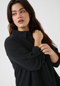 Camisa de botones negra con cuello, mangas enrolladas, textura suave y corte relajado. Diseño minimalista sin patrones.