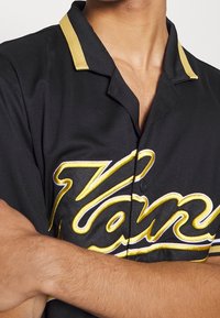 Persona che indossa una camicia nera con bottoni, con accenti dorati e scritte ricamate gialle, con le braccia incrociate sul petto.