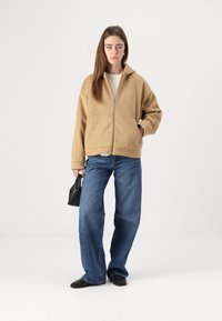 Beige hettejakke med glidelås i fleece, to frontlommer, kombinert med løse blå jeans og svarte flate sko, holdt en svart håndveske i hånden.