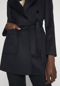 Gros plan sur une personne portant un manteau noir ceinturé avec de grandes poches avant, une main à l'intérieur d'une poche, sur un fond uni.