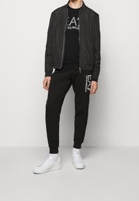 Man som bär svart zip-jacka, svart sweatshirt och svarta sweatpants med vitt EA7 Emporio Armani-logga, vita sneakers och vita strumpor.