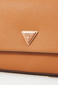Tan-värinen nahkapinta, jossa on kolmion muotoinen Guess USA -metallilogo keskittyneenä taitteen ylle laukussa tai lompakossa.