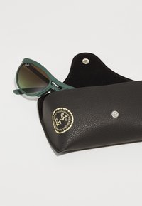 Ochelari de soare Ray-Ban cu ramă mată verde și lentile cu degradé, așezați într-o carcasă din piele texturată neagră, cu un logo auriu.