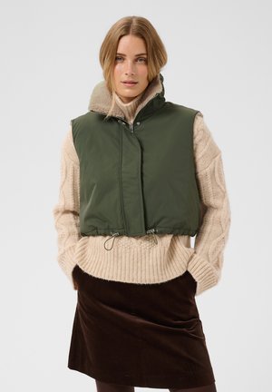 Femme portant une doudoune sans manches vert olive sur un pull beige à torsades et une jupe en velours côtelé marron foncé, debout les mains dans les poches.