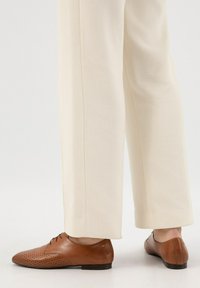 Scarpe eleganti in pelle marrone con design perforato, dettagli marrone chiaro e un tacco piccolo, abbinate a pantaloni a gamba larga color crema.
