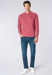 Felpa girocollo bordeaux con polsini a coste, abbinata a jeans blu e sneakers bianche. Design casual e aderente con un piccolo emblema sulla parte anteriore.