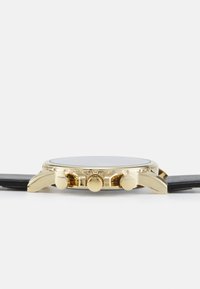 Montre dorée de forme ronde, bracelet en cuir noir et trois poussoirs texturés. Verre lisse avec un design minimaliste.