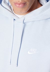 Světlomodrá mikina s měkkou texturou, s bílým logem Nike na hrudi a nastavitelnými šňůrkami na kapuci.