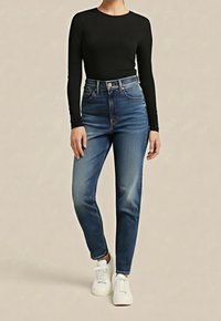 Personne portant un haut noir ajusté à manches longues, un jean bleu taille haute et des baskets blanches, debout devant un fond beige uni.