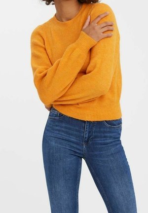 Personne portant un pull à manches longues orange vif et un jean bleu taille haute, bras croisés avec une main sur l'épaule.