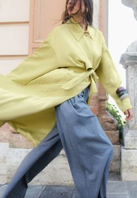 Gelbe, übergroße Jacke mit gebundenem Taillengürtel und einem Kragen mit Knöpfen. Kombiniert mit weiten, grauen Hosen aus einem strukturierten Stoff.