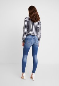 Blå smala jeans med en urblekt textur, kombinerade med en svart och vit randig blus. Modellen bär vita högklackade skor, sedd bakifrån.