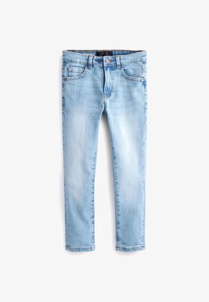 Personne portant un jean slim bleu clair, des baskets blanches et un t-shirt décontracté gris clair, debout devant un fond uni.