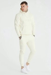 Cream-colored hoodie and joggers made from soft fabric. Features ribbed cuffs and a front pocket. Paired with white sneakers.