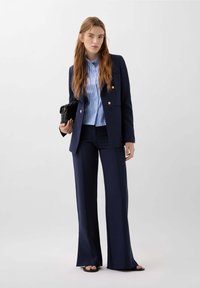 Námořnický modrý tailoring oblek s rozšířenými kalhotami a dvouřadým saku, nošený přes světle modrou pruhovanou košili, drží černou kabelku.