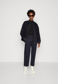Jachetă bomber marină cu manșete răsucite, pulover negru cu guler înalt, pantaloni navy ajustați și pantofi sport albi cu șireturi. Design și texturi minimaliste.