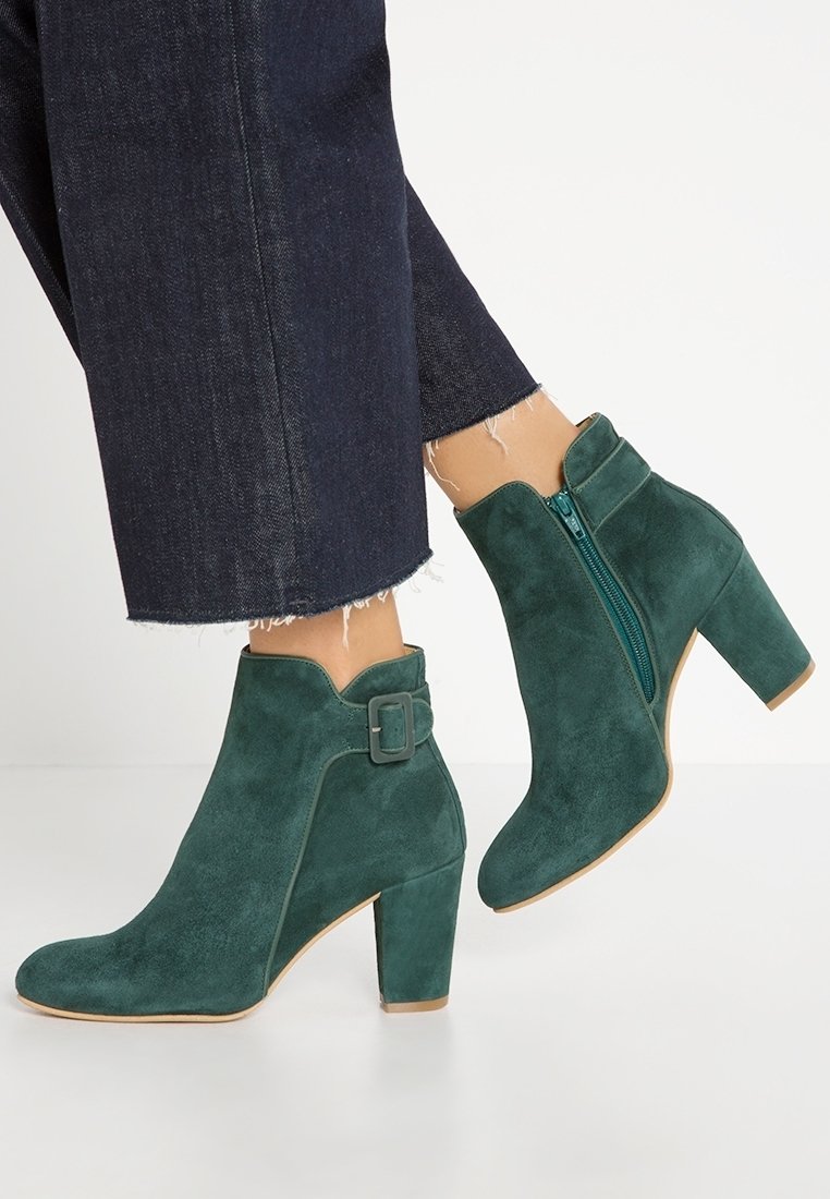 Bottes en daim vertes montant à la cheville avec talons blocs portées avec un jean bleu foncé raccourci et effiloché, montrant une fermeture éclair latérale et une boucle décorative.