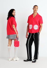 Červená krátkourá zástera s bielymi motívmi sedmokrások, kombinovaná s čiernymi nohavicami s bielym lemom. Model drží rúžovú tašku s volánmi a má na sebe biele pančuchy po kolená.