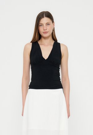 Γυναίκα με μακριά καστανά μαλλιά που φοράει μαύρη αμάνικη μπλούζα με V-λαιμόκοψη και λευκή φούστα, στέκεται μπροστά σε λευκό φόντο.