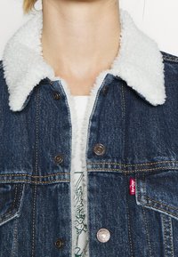 Jeansjacka med en vit fluffig krage, bronsknappar och gula sömnadsdetaljer. Levi's logotyp patch visas på framsidan nedtill.