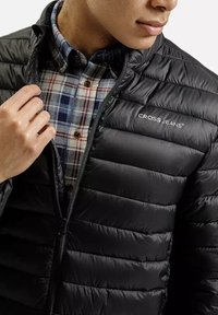 Czarna kurtka puchowa o gładkiej, błyszczącej fakturze i poziomym przeszyciu. Noszona na niebieskiej koszuli flanelowej w kratę z brązowymi guzikami.