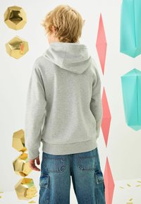Grå hættetrøje med en stor hætte, der har en blød struktur. Sammen med blå denim cargo-bukser. Baggrunden indeholder geometriske dekorationer.