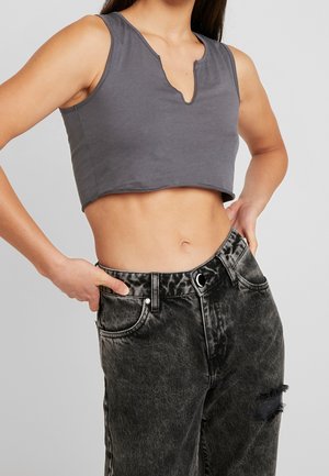 Femme portant un haut court sans manches gris foncé avec un décolleté en V échancré et un jean noir délavé taille haute avec un patch usé.